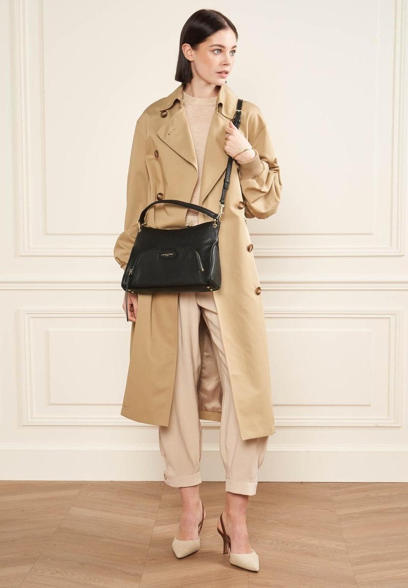Mackintosh beige con grandi bottoni, borsa in pelle nera con dettagli dorati, top color crema e pantaloni chiari abbinati a tacchi beige.