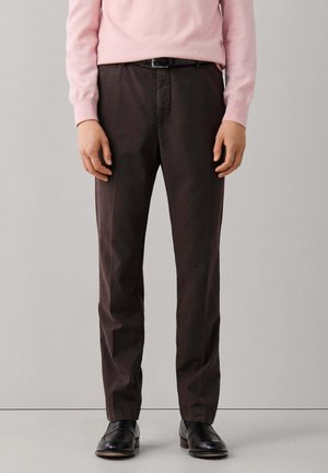 Hombre con pantalones marrón oscuro, cinturón negro, zapatos negros y un suéter de manga larga rosa claro, de pie frente a un fondo liso.