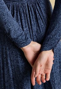 Tumedat, mustriline, pika käistega ja kokkuvõetud vöökoht. Käed on selja taga koos, vasakul käel on näha delikaatne sõrmus.