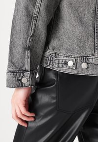 Gray denim jacket with a textured finish, featuring silver snap buttons and a close-up view of the cuff. Black faux leather pants are visible.