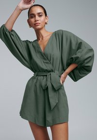Zelené zavinovacie šaty s hlbokým výstrihom do V, nadýchanými rukávmi a opaskom na zaväzovanie. Vyrobené z textúrovanej látky, s uvoľneným strihom a šortkami.