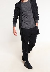 Zwarte lange jas, grijs shirt, versleten zwarte broek en zwarte instappers. Heeft een relaxte pasvorm en een casual ontwerp met zichtbare scheuren.