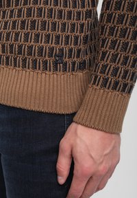 Gestrickter Pullover mit einem geometrischen Muster in Braun und Schwarz, geripptem Saum und Bündchen. Weiche Textur, taillierte Passform und Logo-Detail an der Seite.