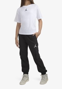 Biała koszulka z krótkim rękawem z czarnym logo Jumpman, połączona z czarnymi spodniami cargo z kieszeniami i elastycznymi mankietami, noszona z jasnymi trampkami.