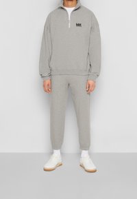 Sudadera gris con cuello alto, media cremallera y detalle de logo. Pantalones deportivos grises a juego con puños elásticos. Zapatillas blancas.