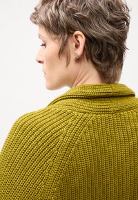 Cardigan verde lavorato a maglia con colletto alto, caratterizzato da un motivo a coste testurizzate e tre cuciture verticali prominenti sul retro.