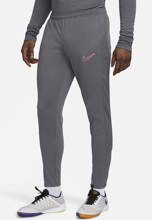 Homme portant un pantalon de sport gris avec le logo Nike rose, des chaussettes blanches et des chaussures de sport d'intérieur blanches avec des lacets noirs et des semelles colorées.