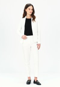 Vit skräddarsydd blazer över en svart topp, kombinerad med vita byxor och svarta loafers med en detalj av guldkedja. Slät tyg, skräddarsydd design.