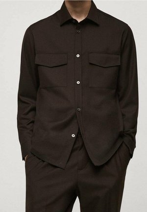 Homme portant une chemise boutonnée marron foncé avec deux poches poitrine, associée à un pantalon marron foncé assorti, mains dans les poches.