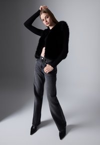 Black faux fur vest over a black long-sleeve top, paired with faded black jeans and pointed black heels; model poses against a gray background.