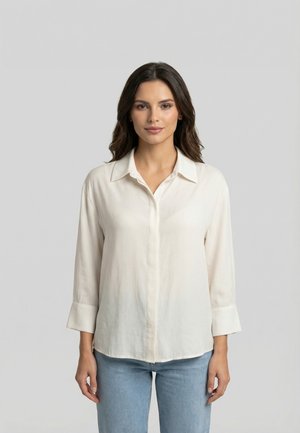 Jeune femme aux cheveux foncés portant une chemise crème à manches longues boutonnée et un jean bleu clair, debout devant un fond gris.