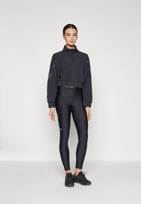 Black cropped jacket with zip and elastic waistband, paired with black fitted leggings. Both feature smooth textures and minimal accents. Black sneakers.