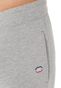 Grå sweatpants med ribbad midja, sidofickor och en liten logotyp i marinblått och rött nära sömmen på fickan. Mjukt tyg.