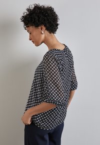 Blouse marine avec un motif géométrique gris, en tissu semi-transparent, col rond et manches trois-quarts, associée à un pantalon foncé.