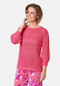 Rosa gestrickter Pullover mit einem lockeren, offenen Gewebemuster. Mit Drei-Viertel-Ärmeln und einem weiten Ausschnitt, kombiniert mit Blumendesign-Hosen.