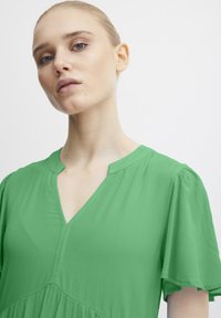 Robe verte à col en V avec de courtes manches volantées, en tissu texturé, et un détail de couture sous la poitrine. L'encolure présente une courbe subtile.