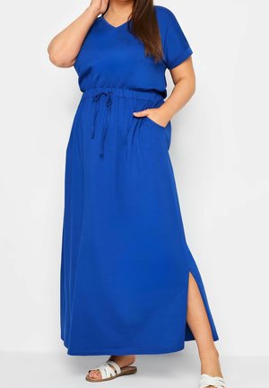 Woman wearing a blue short-sleeve maxi dress with side slit, drawstring waist, and white sandals, hand in pocket and touching hair.