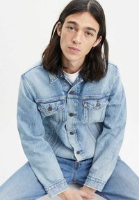 Jasnoniebieska dżinsowa kurtka z dwoma kieszeniami na piersi, zapinana na guziki, z klasycznym kołnierzykiem. Nosi się ją na białej koszulce, w zestawie z jasnoniebieskimi dżinsami.