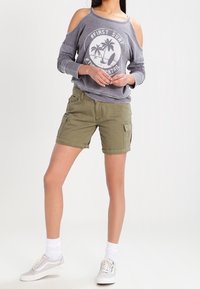 Sweat-shirt gris à épaules dénudées avec un graphisme blanc, short cargo vert olive, et baskets gris clair avec semelles blanches et chaussettes blanches.