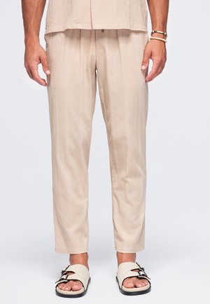Homme portant un pantalon léger beige et un haut assorti, des sandales beiges à double brides, et des bracelets au poignet droit, debout sur fond blanc.