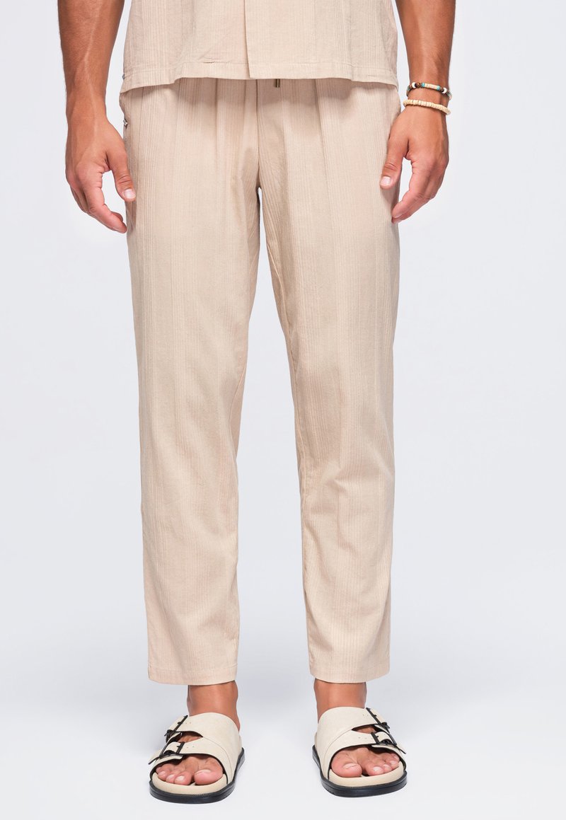 Homme portant un pantalon léger beige et un haut assorti, des sandales beiges à double brides, et des bracelets au poignet droit, debout sur fond blanc.