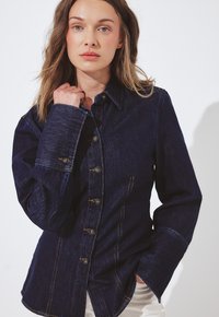 Vrouw met lichtbruin haar, gekleed in een donkerblauwe denimjas met knopen, die de kraag met één hand vasthoudt, voor een plain witte achtergrond.