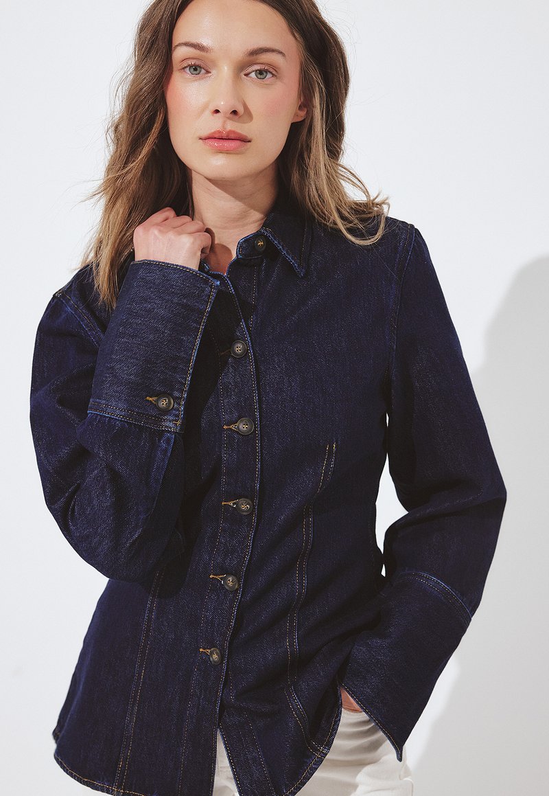 Vrouw met lichtbruin haar, gekleed in een donkerblauwe denimjas met knopen, die de kraag met één hand vasthoudt, voor een plain witte achtergrond.