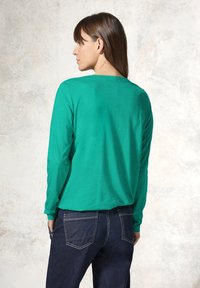 Langärmliges, grünes Jersey-Top mit lässiger Passform, Rundhalsausschnitt und glatter Textur, kombiniert mit dunkelblauen Jeans.