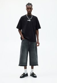 Black oversized t-shirt with logo, gray wide-leg denim shorts with frayed hem, white socks, and black loafers. Silver chain necklace detail.