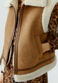 Nahaufnahme einer braunen Wildlederweste mit weißem Schaffellbesatz, silbernem Reißverschluss und seitlicher Tasche, getragen über einem langärmligen Kleid mit Leopardenmuster.