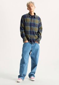 Groene en marineblauwe geruite blouse met knopen en twee borstzakken, gecombineerd met losse blauwe spijkerbroek en sneakers met roze accenten.