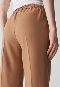 Pantaloni beige con una texture morbida, dotati di una vita arricciata, leggermente svasati e due tasche posteriori. Il tessuto sembra leggero e liscio.