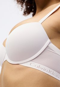 Biustonosz Calvin Klein z miękkiej białej tkaniny, z formowanymi miseczkami, przezroczystymi bocznymi częściami i ramiączkiem z haftowanym logo. Regulowane ramiączka.