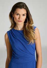 Niebieski top bez rękawów z drapowaniem z przodu, gładka faktura i dopasowana talia. Modelka nosi dużą biżuterię w postaci kolczyków; tło jest neutralne, szare.