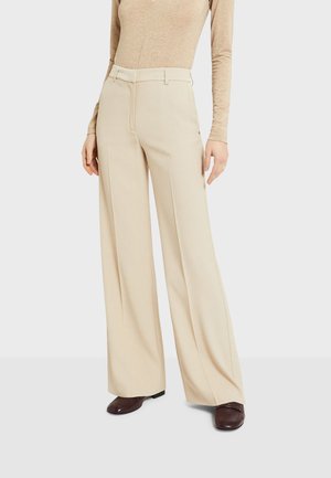 Femme portant un pantalon crème taille haute à jambes larges, associé à un top beige à manches longues et des chaussures marron foncé à bout carré.