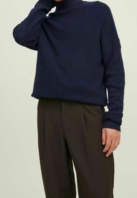 Persona che indossa un maglione a collo alto texturizzato blu navy e pantaloni larghi marrone scuro, in piedi davanti a uno sfondo uniforme.
