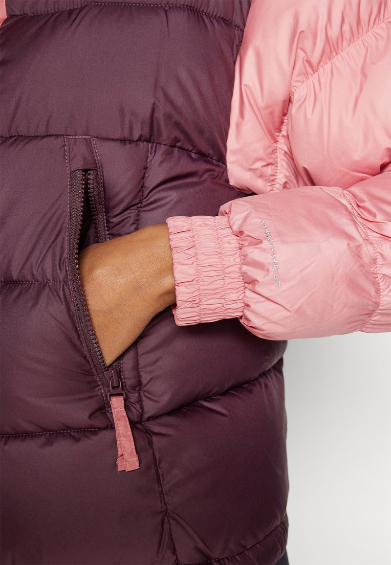 Persona che indossa una giacca rosa con "OMNI-HEAT" sulla manica, con una mano inserita nella tasca con zip di una giacca a vento bordeaux.