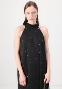 Črna obleka s halter vratnim izrezom, ki se ponaša s tkanino, ki pada v svobodnem slogu, okrašena z manjšimi srebrnimi okrasnimi elementi in teksturirano tkanino.