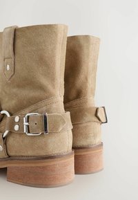 Stivali ankle in suede beige presentano una punta rotonda, un dettaglio con fibbia e un tacco in legno con elementi di design a strati. La texture morbida ne aumenta la durata.