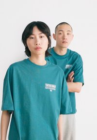 Turquoise oversized t-shirts with short sleeves, featuring a white graphic logo on the front, on two models standing against a white background.