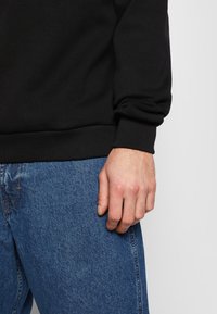 Czarna bluza z prążkowanymi mankietami i dołem, zestawiona z niebieskimi jeansami. Ręka spoczywa na udzie, ukazując fakturę tkaniny obu ubrań.