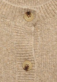 Beige ullkofta med ribbtextur och stor, ljusbrun knapp med fyra hål, som visar en mjuk, stickad konstruktion.