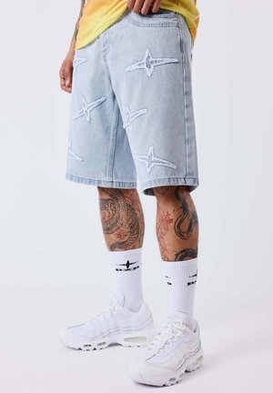 Lichtblauwe denim shorts met stervormige versleten plekken, witte sneakers, witte crew-sokken met zwart logo en getatoeëerde benen zichtbaar tot de knieën.