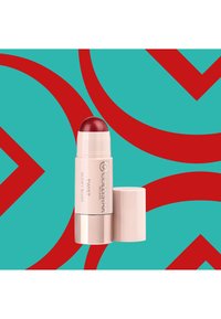 Stick de blush rétractable dans un emballage crème avec une pointe arrondie rouge. L'arrière-plan présente un turquoise avec des formes abstraites rouges audacieuses.