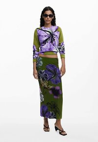 Strikket sweater i grøn med lilla blomsterdesign, kombineret med en lang grøn nederdel med store blomsterprint. Modellen bærer sorte solbriller.