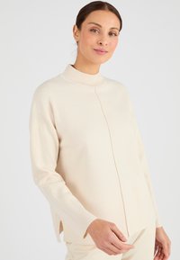 Maglione color crema con collo alto, polsini a coste e spacchi laterali. Tessuto morbido con una vestibilità comoda e dettagli di cucitura frontale.