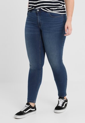 Osoba ubrana w ciemnoniebieskie obcisłe dżinsy, czarno-białe trampki oraz czarno-białą koszulkę w paski, stojąca na jasnoszarej podłodze.