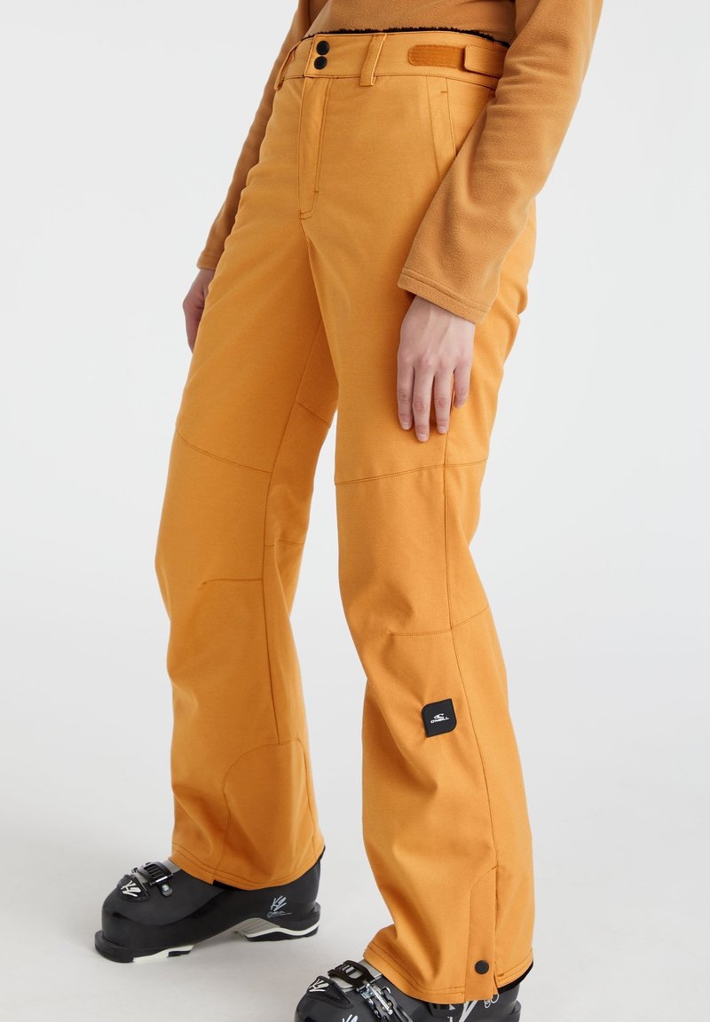 Osoba w musztardowożółtych spodniach i dopasowanej bluzce, stojąca w czarnych butach narciarskich na jednolitym białym tle.