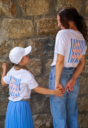 Woman and girl holding hands, wearing matching white "La Dolce Vita" t-shirts, standing against a stone wall, girl in blue skirt and cap.