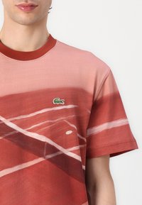 Sarkans grafiskais T-krekls ar mīkstu tekstūru, ar tenisa laukuma dizainu un mazu zaļu Lacoste krokodila logo uz krūšu.